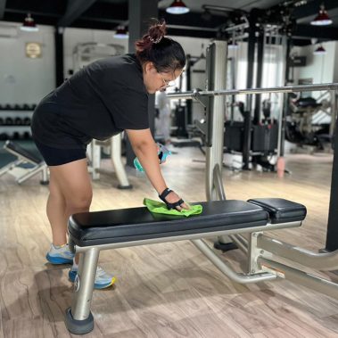 Seorang member wanita di BisaFitness sedang mengelap bench setelah digunakan, menunjukkan sikap disiplin dan menghormati etiket di gym untuk menjaga kebersihan bersama. Foto ini diambil di area latihan yang bersih dan tertata rapi. BisaFitness adalah gym dan fitness center di Batam yang ramah pemula, bersih, dan affordable, dengan dua cabang terdekat di Tiban dan Batam Center.