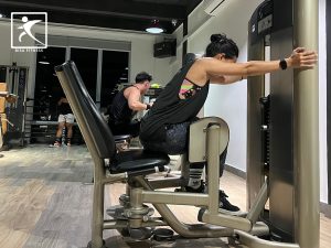 Seorang member di BisaFitness, sebuah gym di Pulau Batam, sedang melakukan latihan dengan abductor machine. Terlihat ia duduk dengan mencondongkan tubuh ke depan, kedua kaki menekan bantalan mesin ke arah luar untuk melatih otot paha bagian luar. Suasana gym terlihat nyaman dengan peralatan kebugaran modern di sekitarnya, memberikan kesan ruang yang bersih dan terawat.