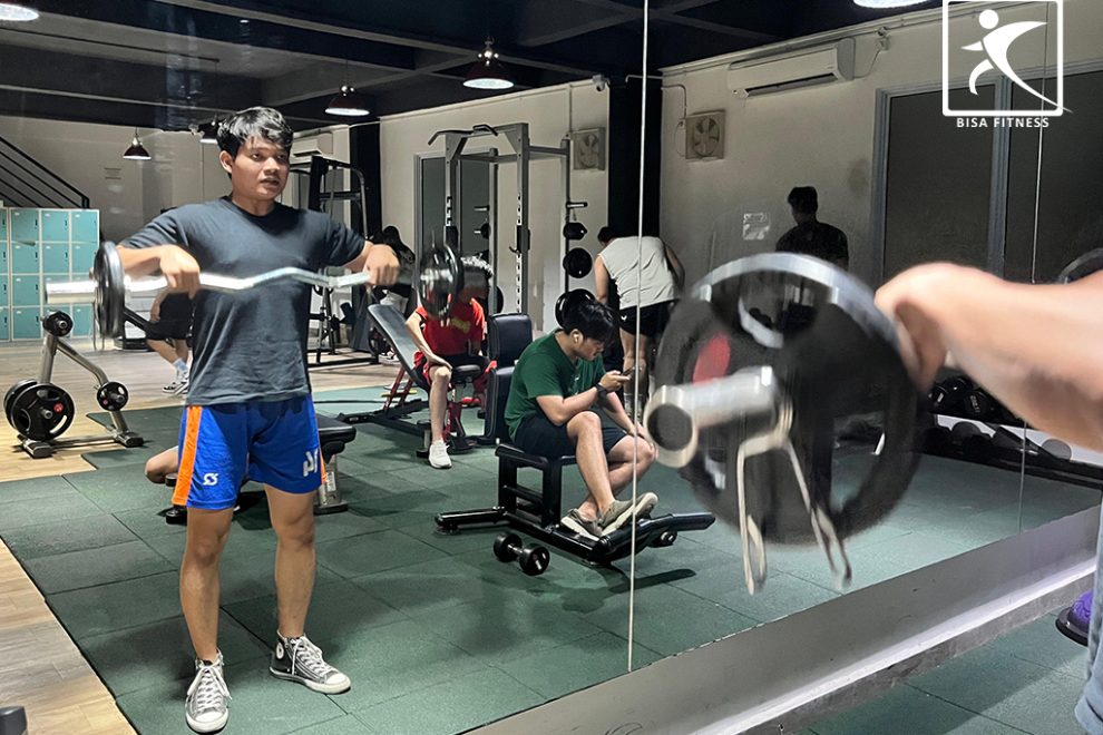 Seorang member di BisaFitness, sebuah gym di Pulau Batam, sedang melatih otot bahu dengan menggunakan EZ Bar melalui gerakan upright row. Ia berdiri tegap dengan kedua tangan memegang EZ Bar, mengangkatnya ke arah dada dengan siku mengarah ke atas. Suasana gym terlihat modern dan teratur, dengan berbagai peralatan kebugaran di sekitarnya, menciptakan lingkungan latihan yang nyaman dan fokus.