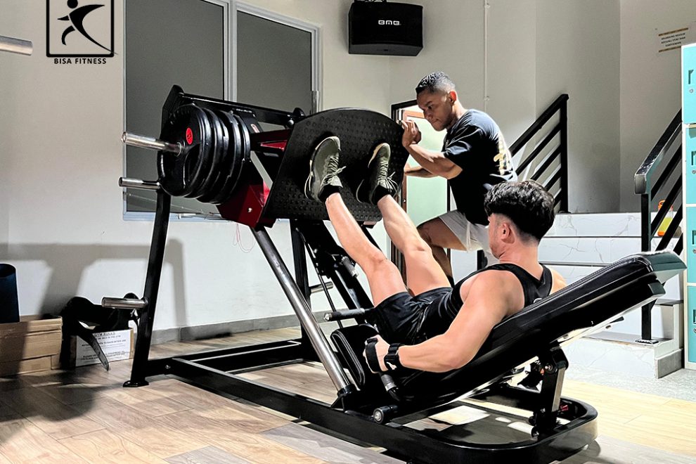 Seorang member di BisaFitness, sebuah gym di Pulau Batam, sedang melakukan latihan menggunakan Leg Press Machine. Ia duduk dengan kaki menekan beban mesin, sementara seorang personal trainer berdiri di samping, memberikan arahan dan memastikan postur yang benar. Suasana gym terlihat dinamis dengan peralatan kebugaran modern di sekitarnya, menciptakan lingkungan latihan yang mendukung dan profesional.