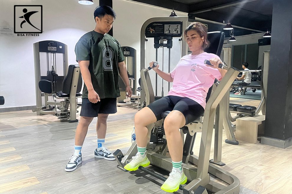 Seorang personal trainer di BisaFitness, gym di Batam, sedang melatih membernya menggunakan Chest Press Machine untuk melatih otot dada. Dengan perhatian penuh, trainer memberikan instruksi dan memastikan member melakukan gerakan dengan teknik yang tepat untuk memaksimalkan hasil latihan. BisaFitness dilengkapi dengan fasilitas modern untuk mendukung berbagai program pelatihan kekuatan dan kebugaran.