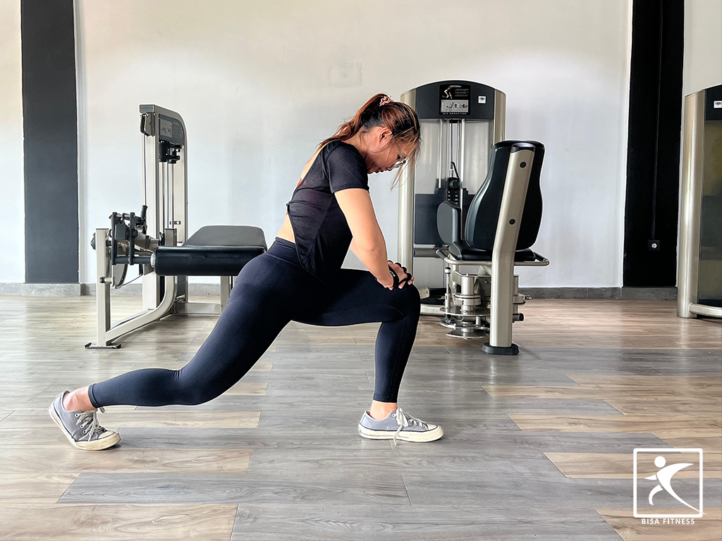 Member BisaFitness sedang melakukan stretching di gym dengan posisi lunges untuk meningkatkan fleksibilitas tubuh, di ruangan yang bersih dan nyaman dengan peralatan olahraga modern. BisaFitness adalah gym dan fitness center di Batam yang ramah untuk pemula, bersih, dan affordable, dengan dua cabang terdekat di Tiban dan Batam Center.