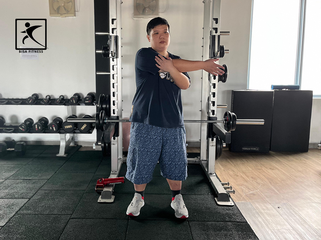 Seorang member pria di BisaFitness tampak melakukan gerakan stretching dengan tangan menyilang di depan dada untuk merilekskan otot bahu dan lengan sebelum memulai latihan. Foto diambil di area gym yang bersih, luas, dan dilengkapi fasilitas lengkap. BisaFitness adalah gym dan fitness center di Batam yang ramah untuk pemula, bersih, dan affordable, dengan dua cabang terdekat di Tiban dan Batam Center.