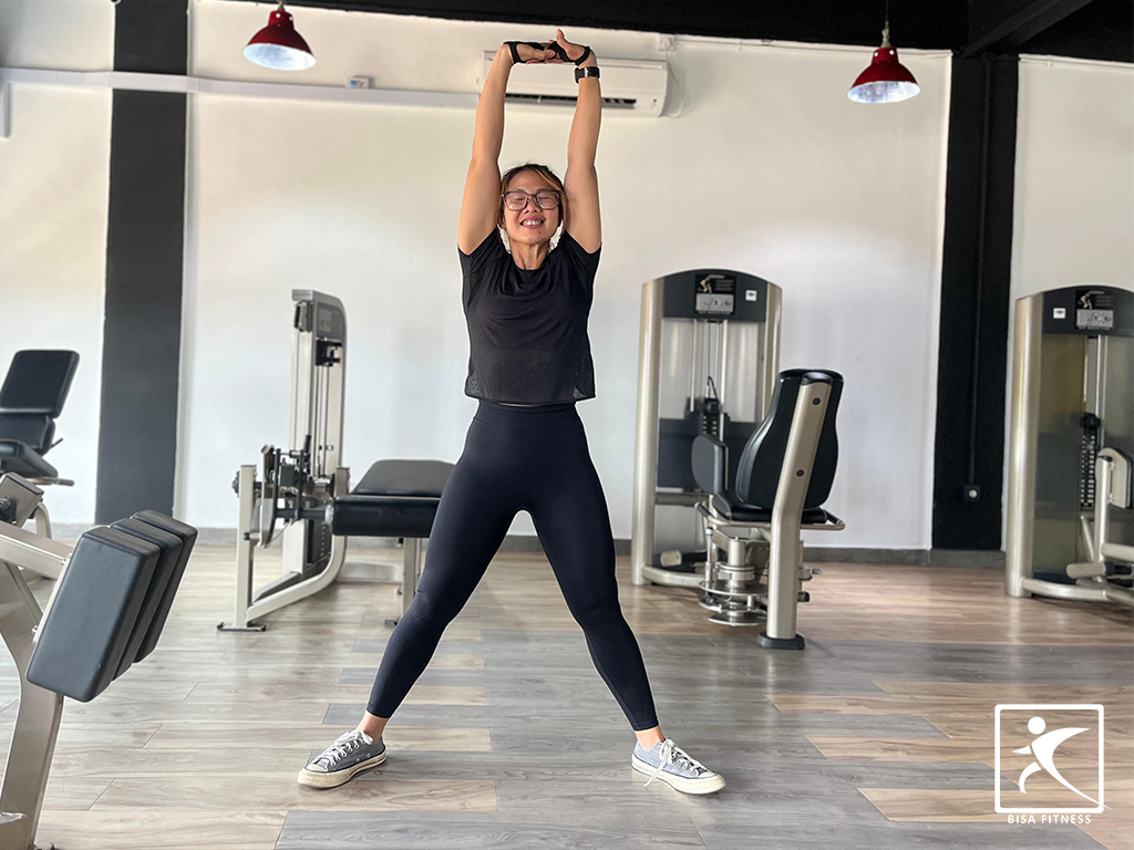 Member BisaFitness melakukan gerakan stretching berdiri dengan tangan terangkat ke atas di ruangan gym yang luas dan bersih untuk membantu meningkatkan fleksibilitas dan mengurangi risiko cedera. BisaFitness adalah gym dan fitness center di Batam yang ramah pemula, bersih, dan affordable, dengan dua cabang terdekat di Tiban dan Batam Center.