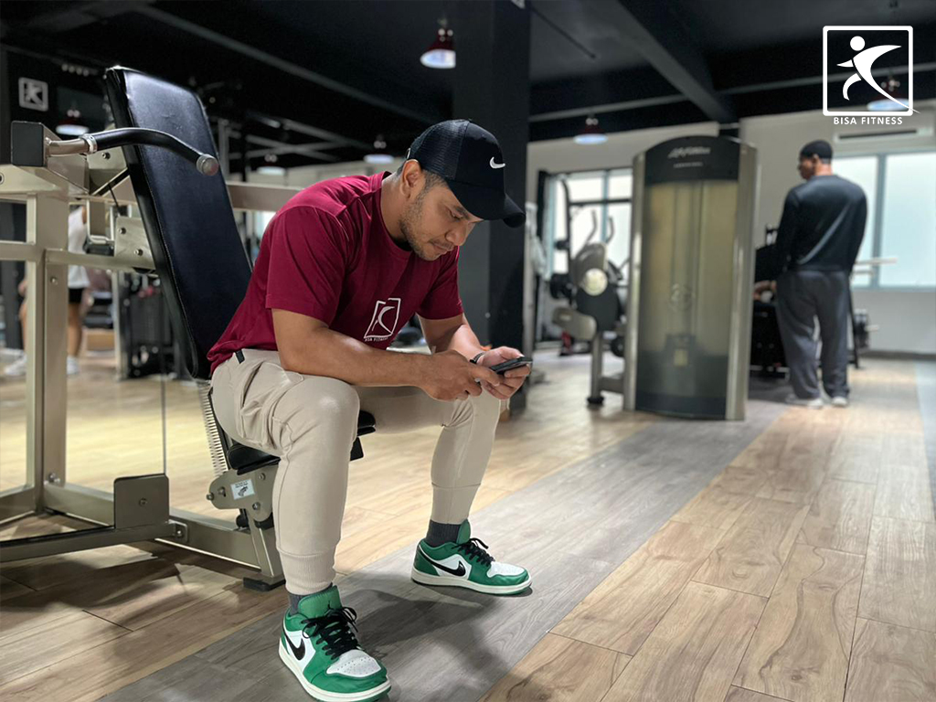 Contoh etika di BisaFitness Batam, gym yang affordable dan pemula-friendly, dengan ilustrasi member duduk di mesin sambil main HP sementara member lain menunggu giliran di cabang Batam Center.